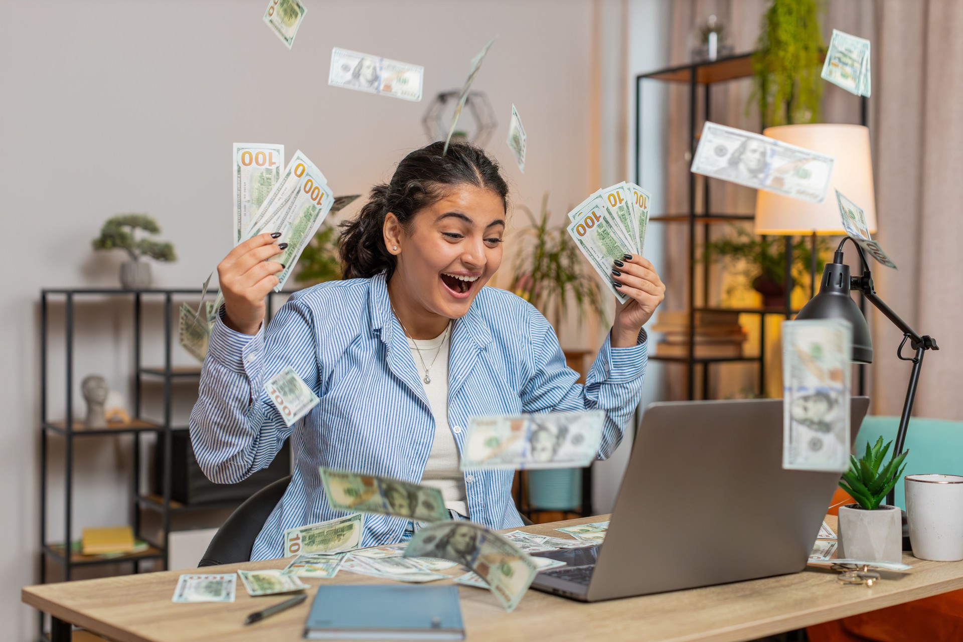 Successful young woman freelancer using laptop celebrate success falling money at home office desk